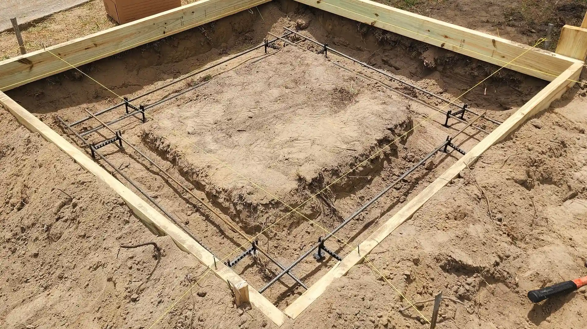 A diamond shaped concrete form with rebar, elevation supports, and string lines set in a dirt foundation.